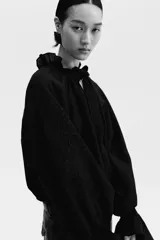 Blusa oversized color negro en tejido liviano de mezcla de lino y viscosa, con cuello elevado plisado y abertura en V con tiras finas. Mangas raglán extralargas con detalles bordados tono sobre tono y puños acampanados con pliegues.