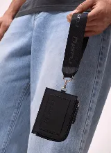 Billetera Rusty modelo Runoi de color negro, con cierre de cremallera y correa de muñeca de tela con el logo de la marca estampado en repetición. Incluye mosquetón metálico plateado para enganchar la correa.