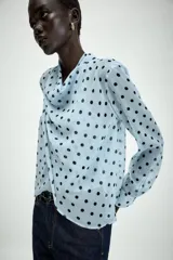 Camisa de manga larga con cuello fluido y diseño cruzado en el frente. Confeccionada en tejido semitransparente de color celeste con estampado de lunares negros. Presenta bajo asimétrico y cierre en la espalda con botones forrados.
