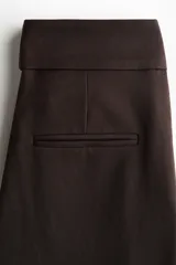Pantalón de sarga color marrón oscuro, de tiro alto y corte bootcut. Cuenta con pretina ancha con detalle de dos botones en el lateral, pliegues marcados en el frente y bolsillos laterales.