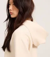 Saco color crema, de corte slim, confeccionado en algodón con viscosa certificada, con cuello con capucha y cordón ajustable, cierre frontal con cremallera y forro de fleece.