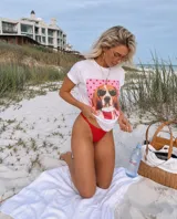Remera blanca de manga corta con estampado frontal de un perro beagle con anteojos de sol sobre un fondo rosa con lunares.