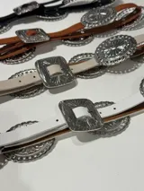 Cinturón marrón de cuero con hebilla plateada rectangular con diseño de flores y apliques metálicos circulares con diseño de flores.