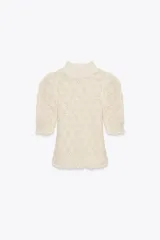 Jersey de punto abierto color crema, con cuello subido y mangas cortas abullonadas. Presenta un patrón de tejido calado con motivos en forma de hoja o escama y un borde festoneado en el ruedo.