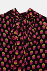 Camisa de cuello subido y manga larga con puños elásticos. Presenta un diseño de lunares en tonos fucsia y amarillo sobre fondo negro, con detalle de pliegues en el frente y cierre de cremallera oculta en la espalda.