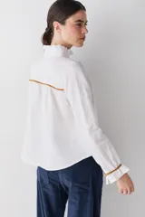 Camisa blanca de lino con cuello alto tipo gorguera, cierre frontal con botones de madera y detalles de ribetes en color mostaza en hombros y puños. Presenta volados en la botonera, cuello y puños.