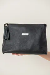 Sobre de cuero negro con textura granulada, cierre superior con cremallera y tirador con borla de flecos. Incluye placa metálica plateada con el logo de la marca.