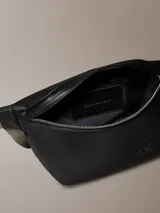 Riñonera Calvin Klein negra de cuero sintético texturizado, con cierre principal y logo CK grabado en el frente. La correa es ajustable y de color gris, con el nombre de la marca repetido.