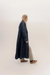 Tapado largo de jean azul oscuro, con cuello solapa y puños con detalle de cinta de terciopelo negro. Presenta costuras visibles que definen paneles y un logo bordado en la espalda.