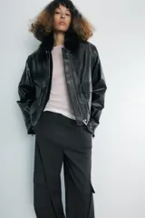 Chaqueta de efecto piel en color negro, con cuello de solapa con aplicación de pelo sintético, manga larga y bolsillos frontales de plastrón con solapa. Cierre frontal con cremallera y botones ocultos bajo solapa.