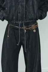 Cinturón de múltiples cadenas metálicas en tonos dorado envejecido y plateado. Presenta secciones de malla con diseño de rombos y dijes colgantes con grabados oscurecidos.