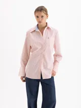 Camisa de corte holgado confeccionada en poplín de algodón, con diseño de rayas verticales en tonos rosados y blancos. Presenta cuello clásico con interior en tela a contratono azul, cierre frontal con botones al tono, mangas largas con puños anchos y un bolsillo plaque en el pecho con detalle de remache metálico en forma de corazón.