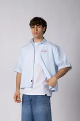 Camisa de manga corta de popelina a rayas celestes y blancas, de corte boxy y calce holgado, con cuello clásico, cierre frontal de botones y bordado en el pecho.