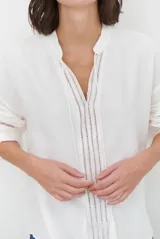 Camisa de color crudo con diseño romántico, cuello redondo con cordón ajustable terminado en borlas, y mangas tres cuartos abullonadas. Presenta detalles de bordados calados verticales en el frente y horizontales en las mangas.