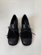 Mocasín de cuero marrón con textura granulada, diseño de horma cuadrada, detalle de flecos y lazo decorativo en el empeine, montado sobre una suela y taco bajo de color negro.