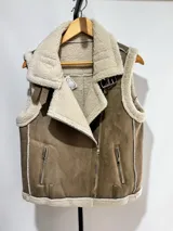 Chaleco estilo gamulán de gamuza color marrón con interior y bordes de corderito sintético color crema. Presenta cierre frontal metálico, bolsillos laterales con cierre y detalles de tiras de cuero sintético en la cintura.