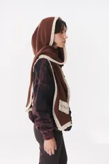 Bufanda de punto color marrón con capucha integrada y bordes en color crema. Presenta una etiqueta rectangular bordada con la palabra Anthea en uno de sus extremos.