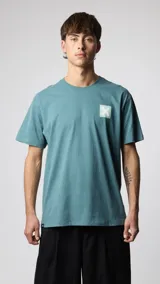 Camiseta de manga corta color verde azulado, corte holgado, con un pequeño estampado cuadrado con una 'X' en el pecho.