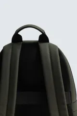 Mochila de color verde militar con textura grabada, diseño minimalista y forma redondeada. Cuenta con un compartimento principal y un bolsillo frontal, ambos con cierre de cremallera. Posee asa de mano y asas de hombro acolchadas y ajustables.
