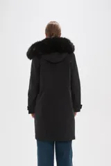 Campera parka negra de corte largo y holgado, con capucha fija adornada con piel sintética, cierre frontal oculto y bolsillos laterales con cordones ajustables en la cintura.