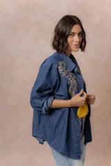 Camisa de denim azul con bordados florales en tonos amarillos y celestes sobre los hombros y el pecho. Presenta cuello clásico, cierre frontal con botones y mangas largas con puños.