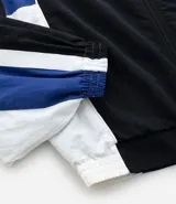 Campera deportiva de poliamida con diseño de bloques de color en negro, blanco y violeta. Presenta cuello alto, cierre frontal con cremallera, puños y ruedo elásticos, y forro interior de poliéster.