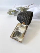 Cinto ancho de cuero negro con textura símil piel de cocodrilo y hebilla rectangular con aplique de guampa en tonos blanco, gris y marrón, con marco plateado.