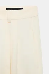 Pantalón wide leg de tiro medio confeccionado en hilatura de algodón, color beige, con trabillas en la cintura, bolsillos laterales ocultos y detalle de pliegues en el delantero. Cierre frontal con cremallera, botón y ganchos metálicos.
