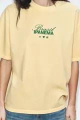 Camiseta amarilla de corte oversize, con cuello redondo, manga corta y estampa frontal en color verde con texto alusivo a Brasil Ipanema y pequeños símbolos.