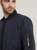 Campera estilo bomber acolchada de color azul marino, con patrón de costuras en rombos. Presenta cierre frontal de cremallera, cuello, puños y dobladillo en punto canalé oscuro. Incluye bolsillo utilitario con cremallera en la manga izquierda con cinta con el logo de Calvin Klein.