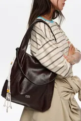 Bolso de mano y hombro grande de cuero marrón oscuro, tipo hobo, con diseño estructurado mediante costuras frontales y asa integrada. Incluye detalle lateral de cordones blancos con cuentas decorativas y el logotipo plateado de la marca en la parte inferior frontal.