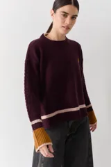 Buzo de punto color crudo con mangas de textura trenzada, detalles en contraste en los puños y franjas horizontales en el ruedo. Presenta cuello redondo y un pequeño bordado en el pecho.