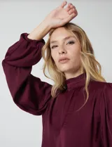 Blusa Tahari de gasa satén color violeta oscuro, con cuello alto fruncido y mangas largas abullonadas. La espalda tiene una abertura central con cierre de botón.