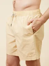 Short de baño tipo volley color amarillo claro, con cintura elástica y cordón ajustable. Presenta un diseño liso con un pequeño logo bordado en la parte inferior de la pierna izquierda.
