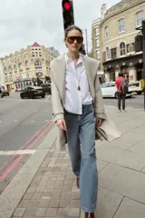 Abrigo de paño negro con capucha, cierre frontal cruzado con botones y cinturón de lazada en el mismo tejido. Presenta manga larga con trabillas en los puños y bolsillos laterales.