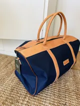 Bolso de viaje tipo duffel de lona negra con detalles en cuero tostado, asas de mano y correa ajustable para el hombro.