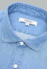 Camisa de jean azul con botones blancos, mangas largas con puños doblados y corte clásico.