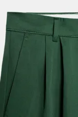 Bermuda de tiro medio color verde oscuro, con trabillas en la cintura, bolsillos laterales y detalle de pliegues delanteros. Presenta un bajo con acabado asimétrico y cierre frontal con cremallera, botón interior y ganchos metálicos. Colección especial Fruit of the Loom con Zara.