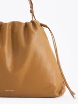 Cartera tipo shopper de cuero vacuno color tostado, con formato amplio, fruncido superior y cordones ajustables a los lados. Cuenta con tiras finas para llevar al hombro y sutil grabado de la marca en el frente.