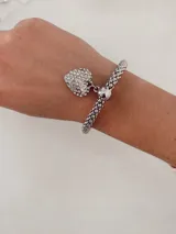 Pulsera elástica de eslabones texturizados en color plateado, con un dije colgante en forma de corazón cubierto de cristales brillantes.