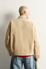 Cazadora relaxed fit color tostado confeccionada en tejido de algodón, con cuello solapa, bolsillos de vivo en la cadera y cierre frontal de cremallera oculta por solapa con alamares metálicos.