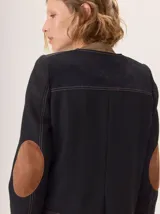 Chaqueta de paño color marrón oscuro, de corte clásico y cuello redondo, con cierre frontal dorado. Presenta bolsillos rectangulares con ribetes en color rojizo y parches de pana a rayas en los hombros.