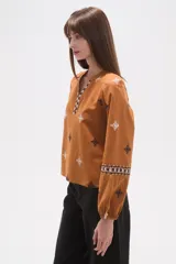 Blusa de algodón color tostado con cuello mao y escote en V. Presenta mangas largas abullonadas con puños elásticos y un diseño con bordados geométricos en contraste distribuidos por toda la prenda.