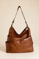 Cartera de cuero natural en color marrón, con diseño envolvente, caída suave y bolsillo exterior cruzado con el nombre de la marca bordado. Cuenta con tiras de hombro, herrajes metálicos y un dije colgante geométrico.