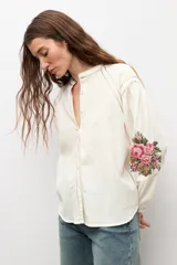 Camisa de color natural con mangas largas abullonadas y bordado floral multicolor en los puños. Presenta cuello mao y cierre frontal con botones.