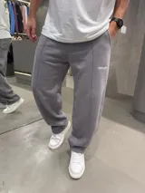 Pantalón deportivo de corte baggy y ajuste holgado, color gris oscuro. Presenta costuras verticales frontales que recorren las piernas y un pequeño logo bordado en la parte superior. Cuenta con bolsillos laterales y cintura elástica.
