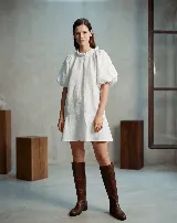 Vestido corto de lino, color blanco, con cuello redondo fruncido, mangas cortas abullonadas y corte evasé con volado en el ruedo.