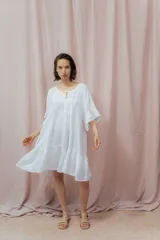 Vestido corto blanco de lino, con diseño suelto y holgado, mangas amplias hasta los codos, escote redondo con abertura en V y lazo decorativo, y un volante en la parte inferior.