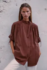 Camisa bordó de cuello alto ajustable con doble hebilla y cierre frontal con botones ocultos por solapa. Tiene mangas cortas con trabillas y diseño holgado.
