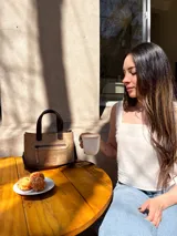 Cartera tipo tote de cuero color beige, con doble asa corta marrón y cierre superior con cremallera. Tiene un bolsillo frontal con cremallera y forro interior de tela con un bolsillo.